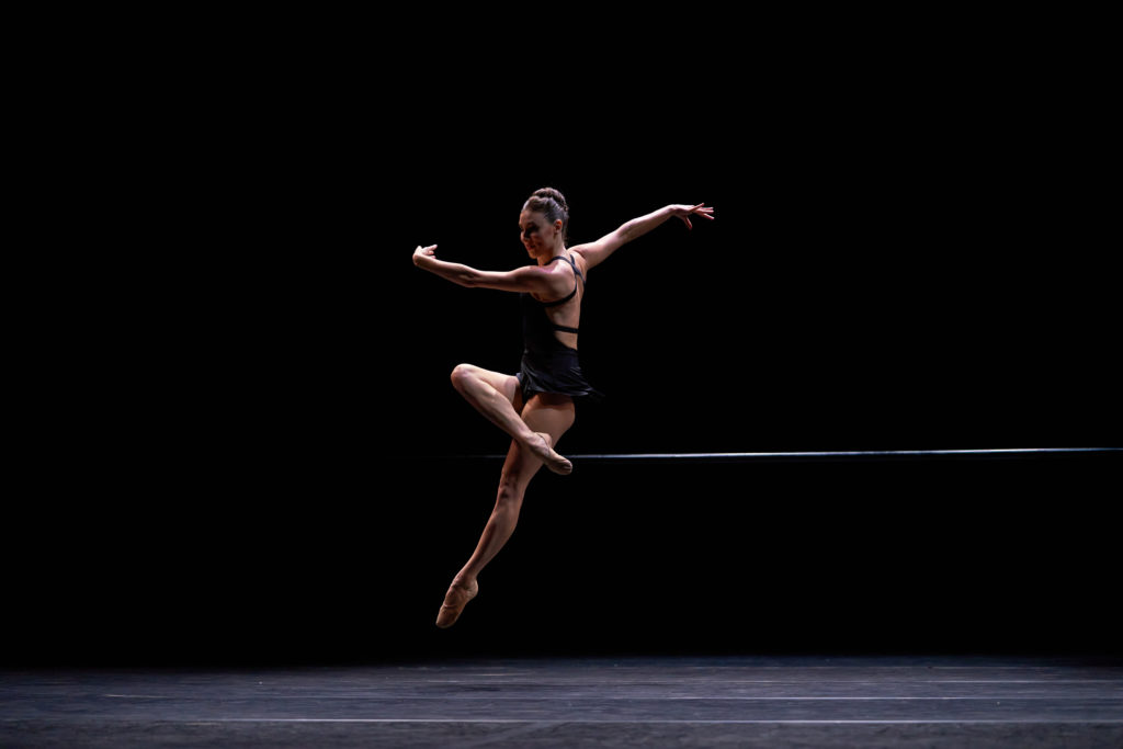 female dancer on darkly lit stage doing a jump with right leg in front attitude.