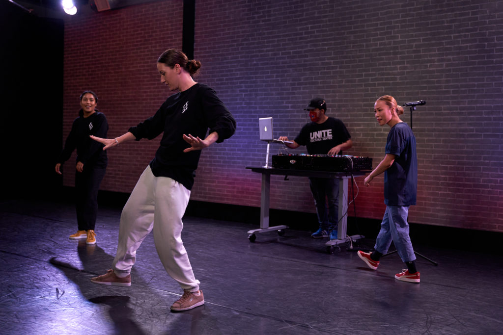 female dancer from femme fatale wearing a black long-sleeved shirt and white sweatpants dancing in front of a DJ