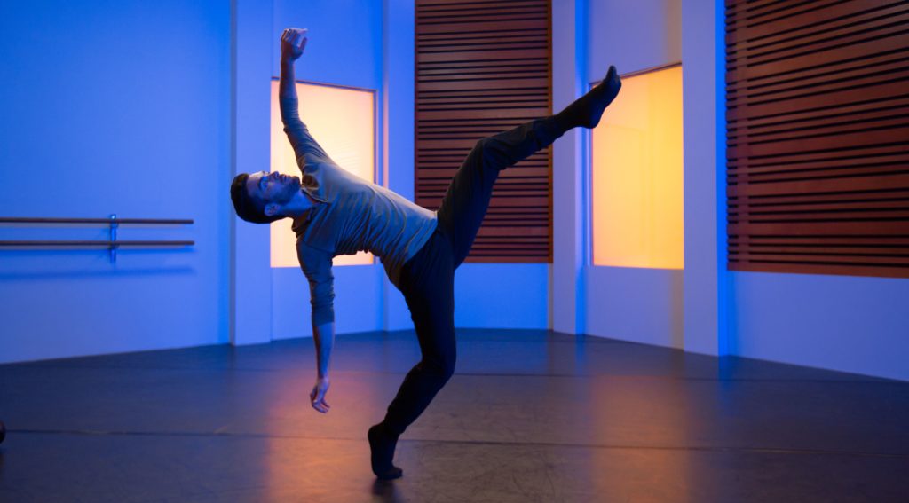 dancer teddy forance in a long sleeved grey shirt and black pants dancing in a blue studio