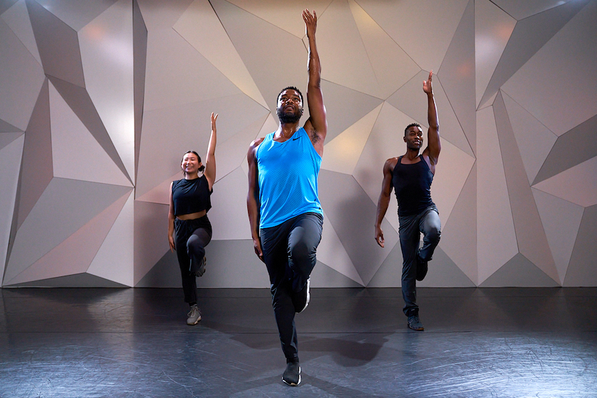 dance instructor grasan kingsberry wearing a blue tank top and black pants standing with one leg up and one arm reaching to the sky 