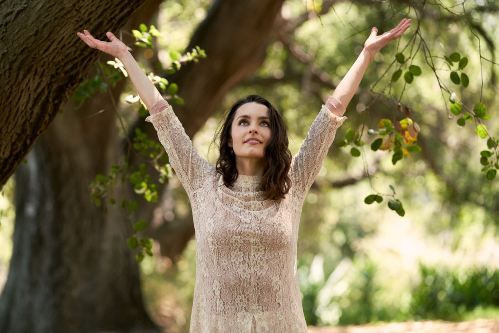 dancer kathryn mccormick wearing a white lace top standing outside and reaching towards the sky while smiling 