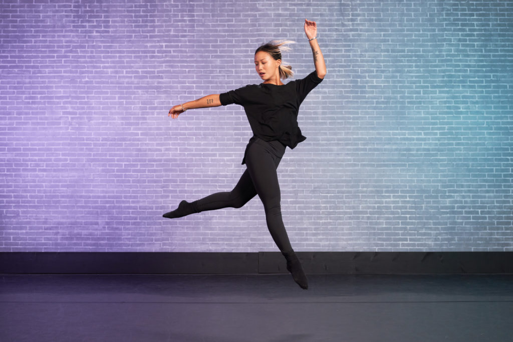 mel mah doing a jump looking over right shoulder infront of blue and purple brick wall at cli studios