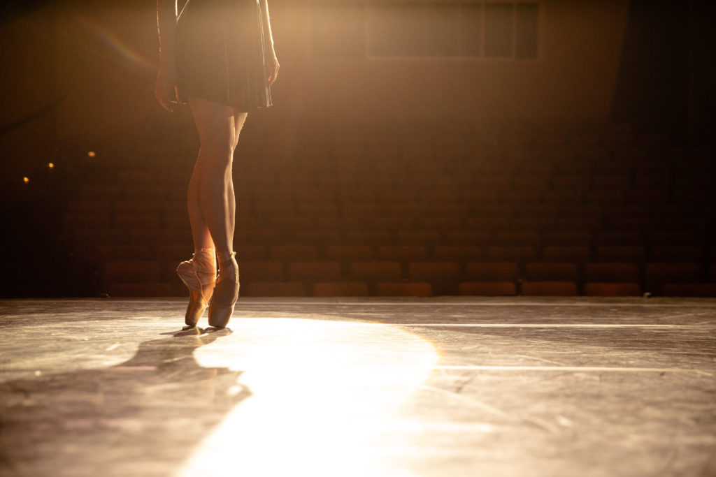 view of tiler peck from waist down from behind on pointe spotlit on stage
