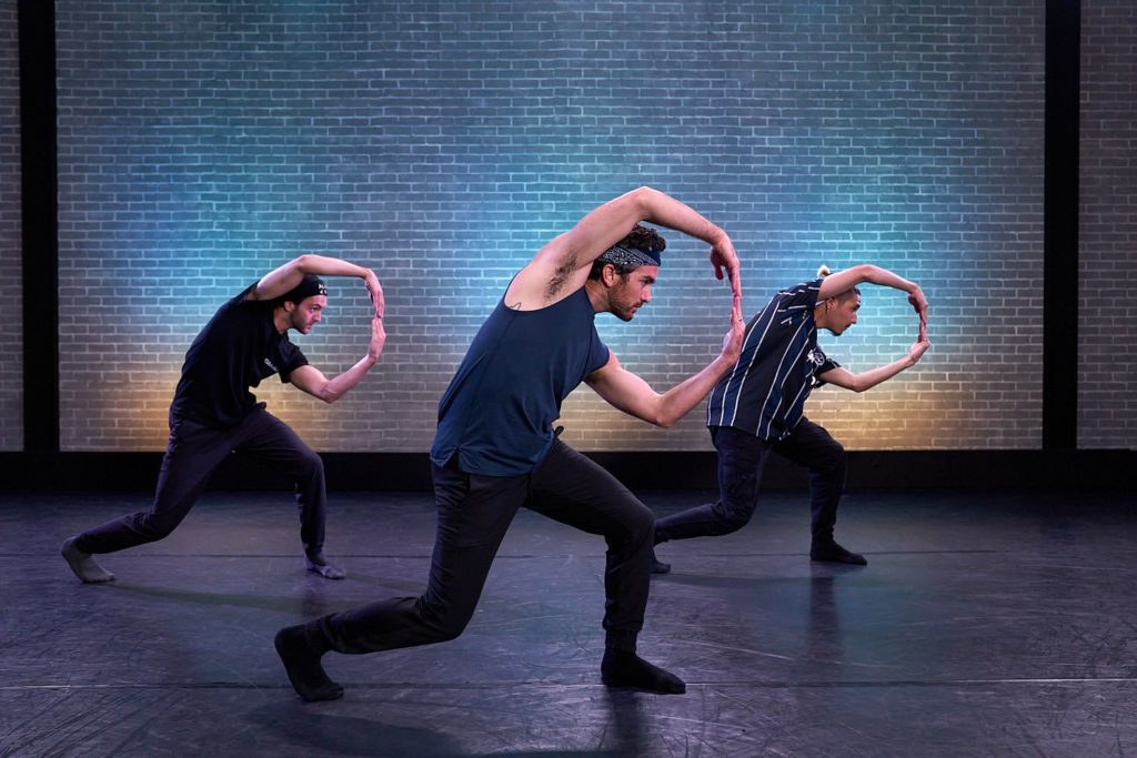 dancer teddy forance and assistants in front of a blue brick wall making a circle shape with their arms and lunging to the side