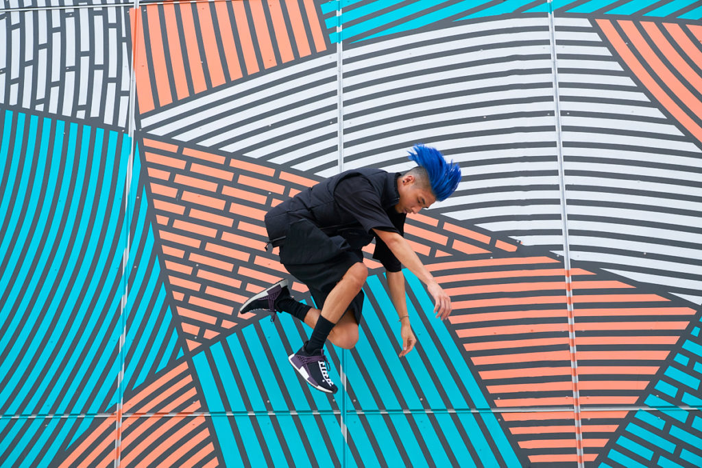 dancer lex ishimoto jumping in front of a blue and orange mural with blue hair and knees tucked in.
