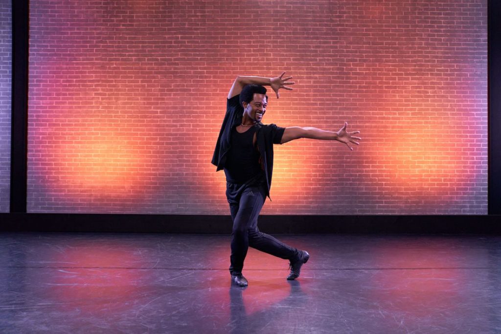 dancer brandon oneal wearing all black striking a jazz pose in front of an orange brick wall