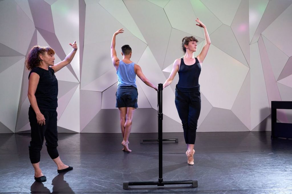 two dancers standing at the ballet barre in relevé with one arm in 5th position 