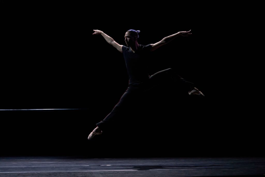 male dancer in all black jumping on a dimly lit stage