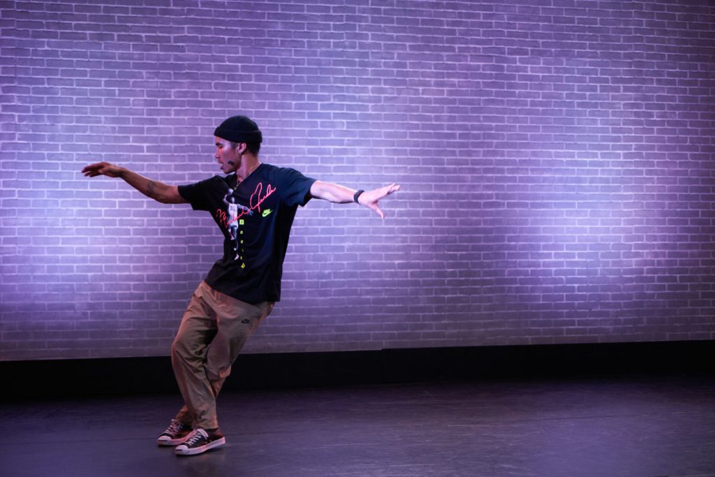 Male hip-hop dancer wearing a black shirt in front of a purple brick wall