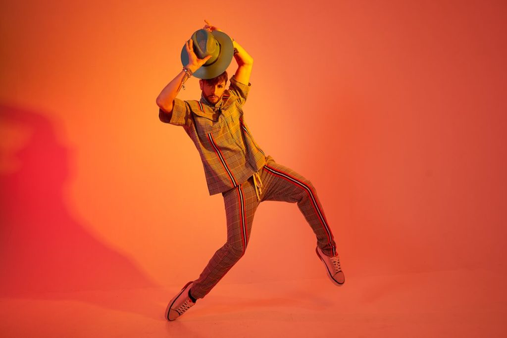 choreographer brian friedman wearing a plaid outfit posing with a hat in front of an orange background