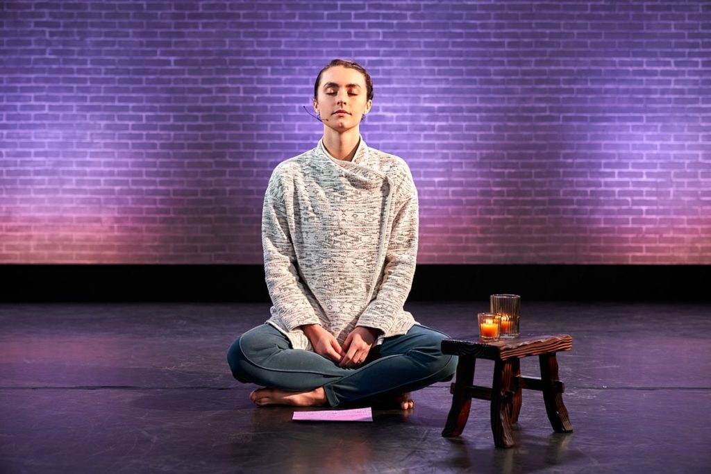 Kathryn McCormick sitting criss crossed in front of the purple wall meditating. 