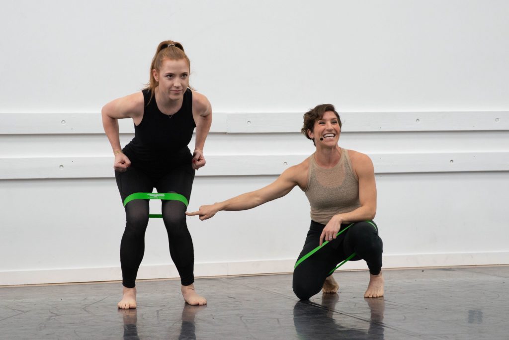 sugarfoot therapy trainer katie scharr pointing to a young dancer who is in a chair position with a strengthening band around her thighs