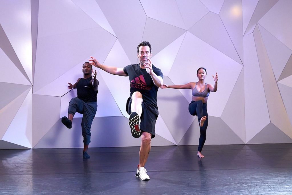 choreographer mark meismer wearing a tshirt and shorts teaching a combination to a female dancer in a sports bra and leggings and a male dancer in joggers and a tank top