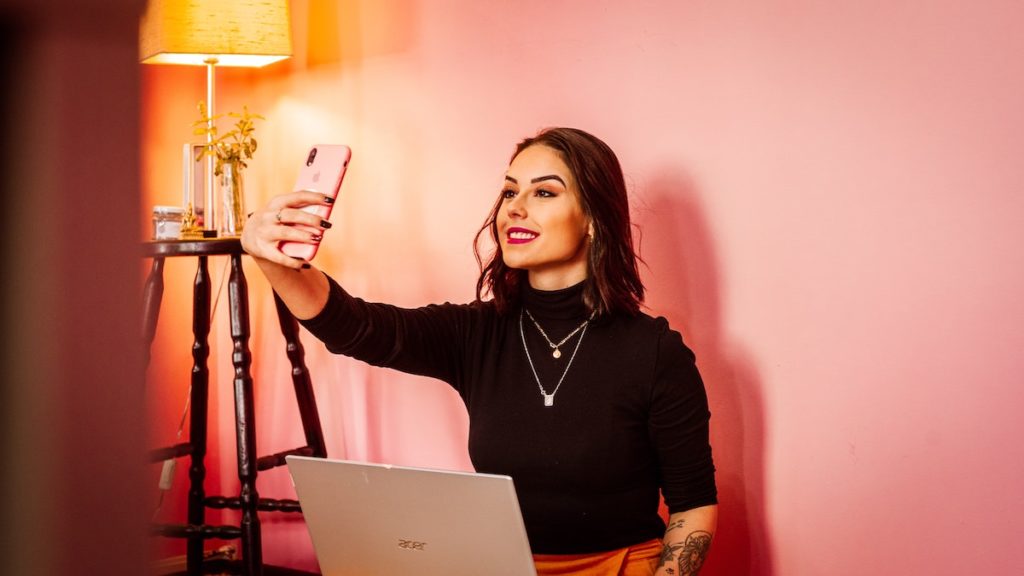 young woman holding an iphone taking a selfie in front of a pink wall 