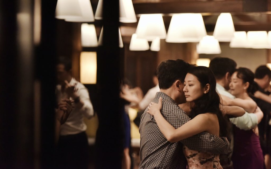 couple embracing and dancing together during a ballroom class with other dancing couples in the background.