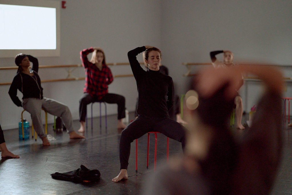 Dancers sitting on stools with their hands on their heads