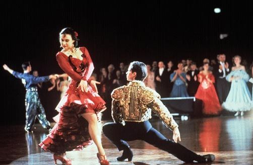 Strictly Ballroom the move with women in red dress and heels standing over man kneeling down dressed in gold costume. 