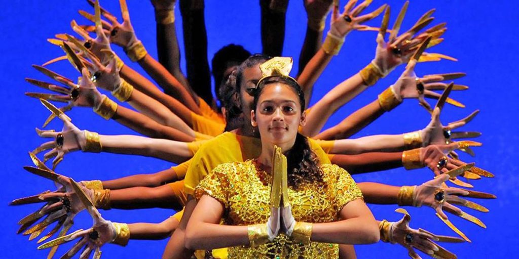 dancer in yellow on blue backdrop with many hands in a circle

