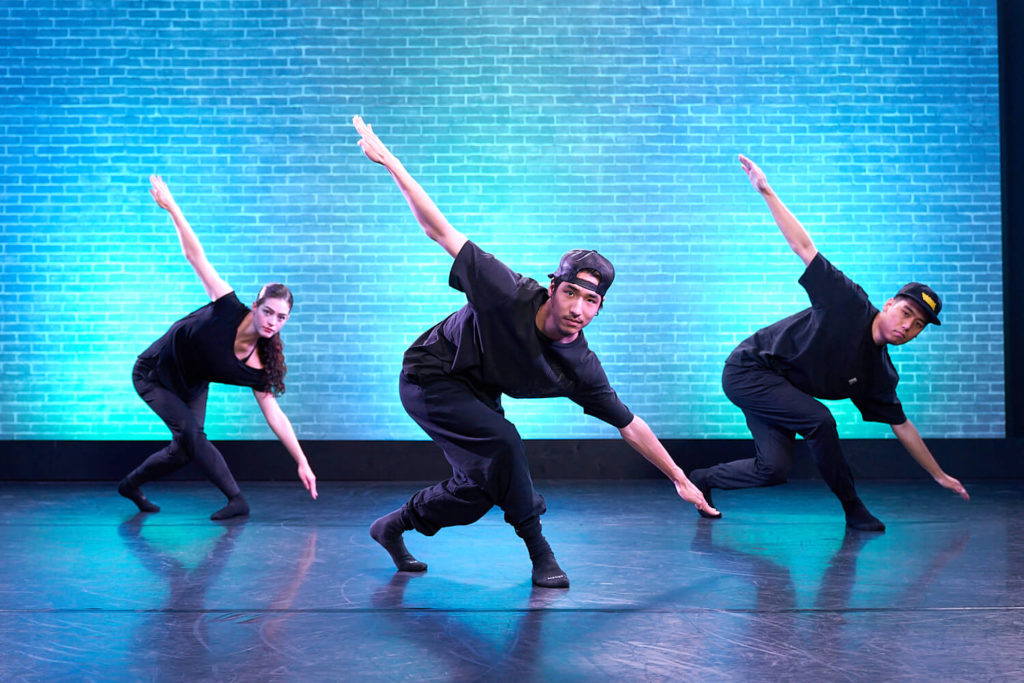 Three dancers with arms straightened in air plane position in front of a blue wall at cli studios.