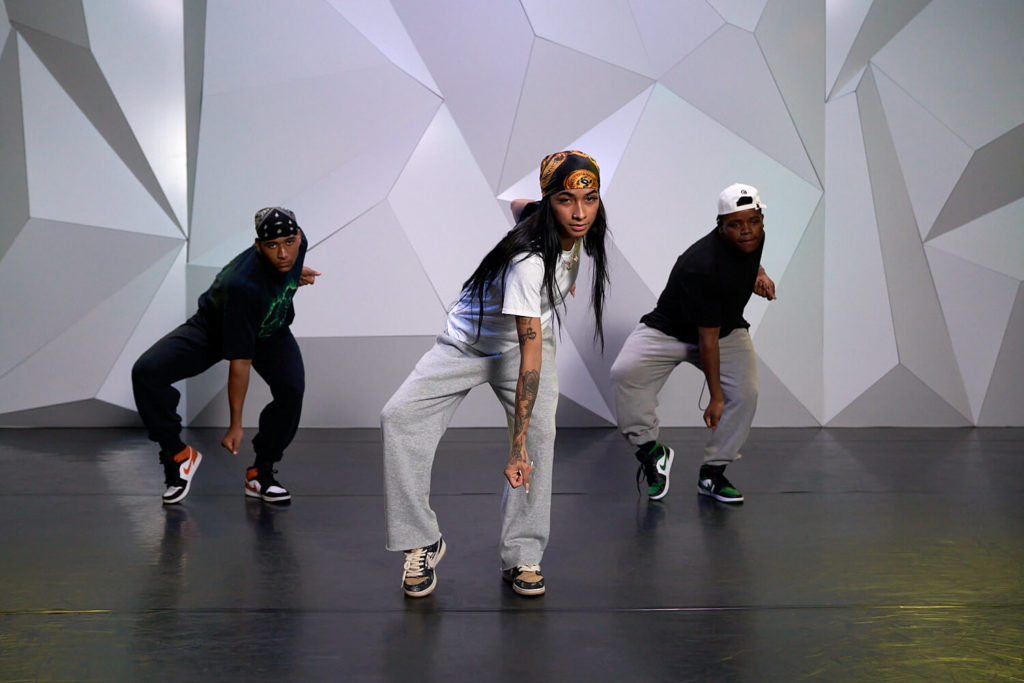 Three female dancers posing in front of a gray wall at CLI studios with arms in a snap position pointing down.