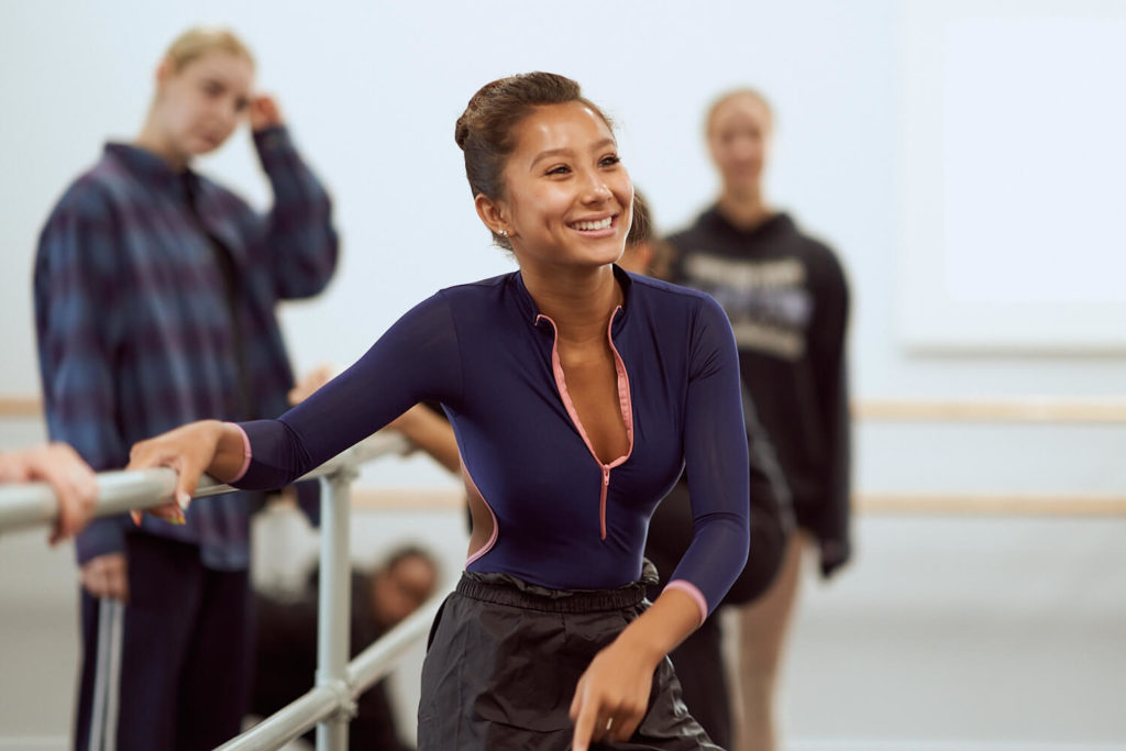 Dancer smiling at a ballet barre wearing a dark blue long sleeved leotard