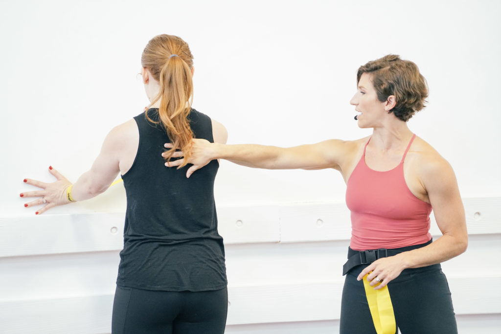 Woman in exercise gear adjusting a woman in black shirt facing a wall