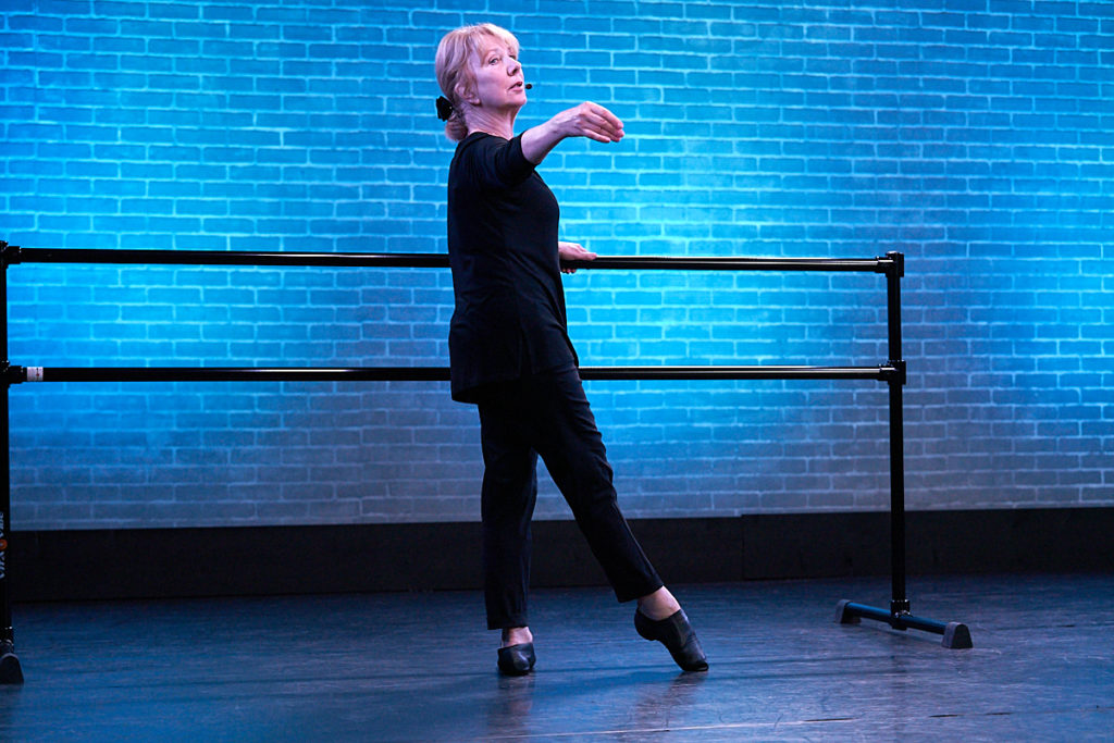 Female Dance teacher dressed in black at the ballet barre with toe in tendu.