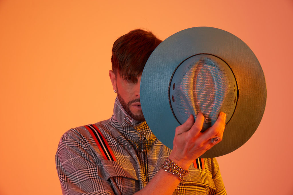 Brian Friedman covers half of face with hat. Orange background.