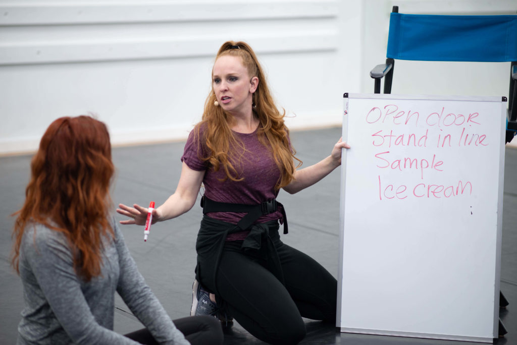 Julie kay sitting on ground in dance studio writing on whiteboard with woman with red hair to her left looking at whiteboard