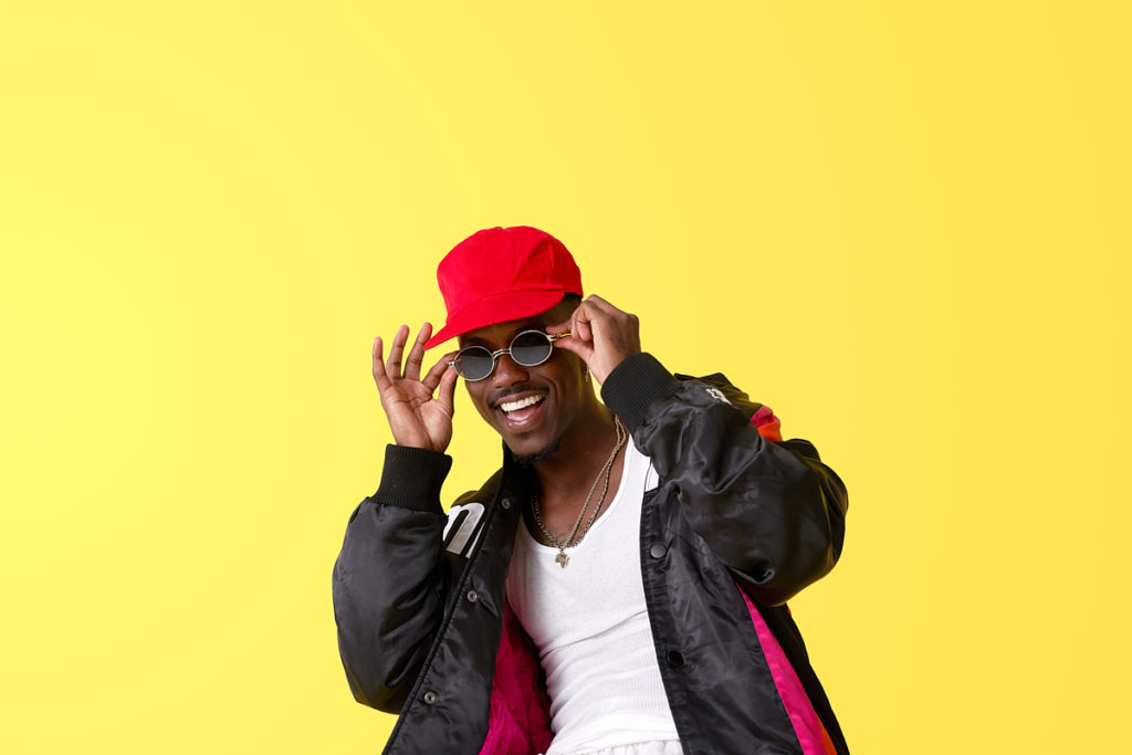 JBlaze headshot wearing red hat in front of yellow background smiling holding his sunglasses to his face