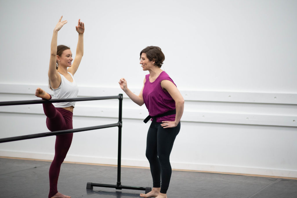 Dancer in red leggings stretching at ballet barre while being coached