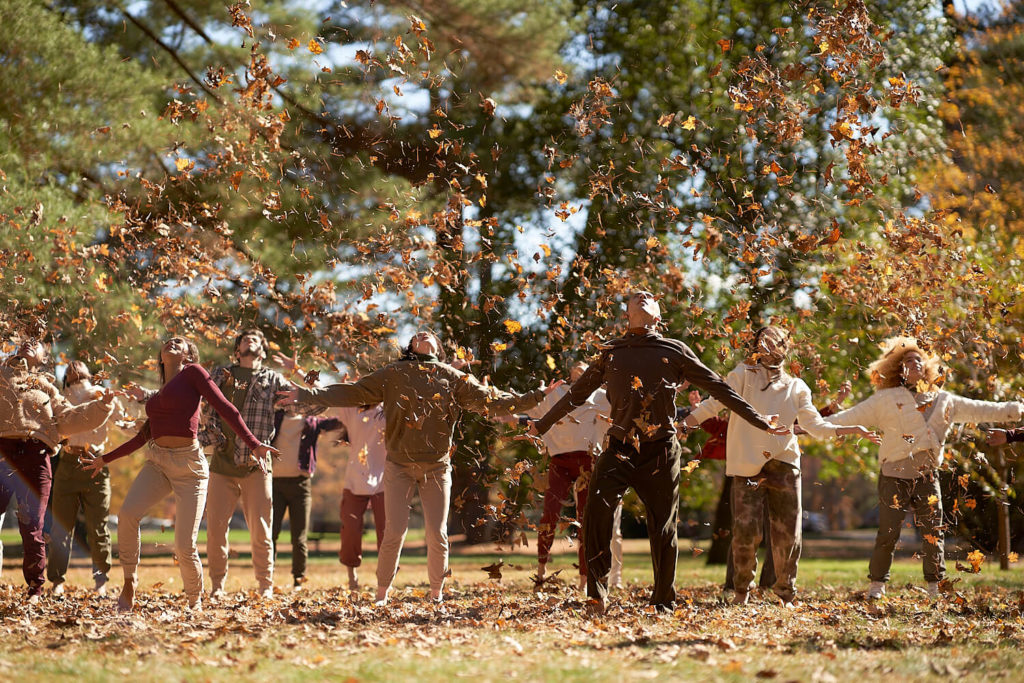 Cli conservatory dancers in a line outside throwing leaves into the air and looking up with arms by their sides.