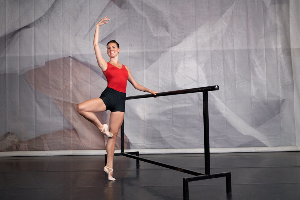 tiler peck at ballet barre with leg in high passé and arm in fifth position in red leotard