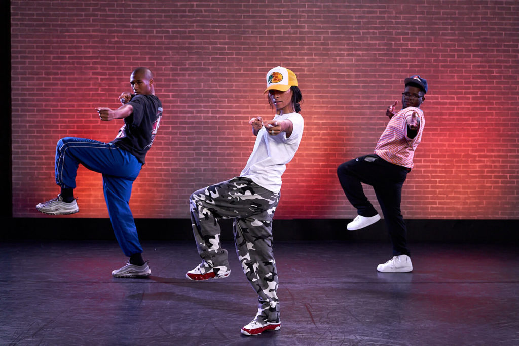 Three hip hop dancers with left leg bent in front of red brick wall projection at CLI Studios