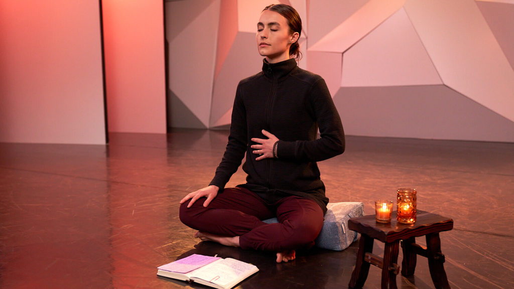 CLI Instructor Kathryn McCormick sitting cross legged on CLI dance studio floor with her hand on her stomach. Meditating with a candle and a notebook
