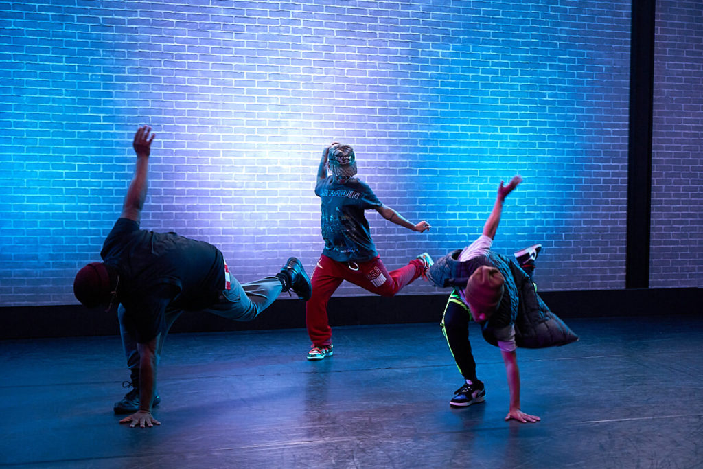 Three hip hop dancers in sweatpants, sneakers and hats touch the ground with one leg up dancing in CLI Studios