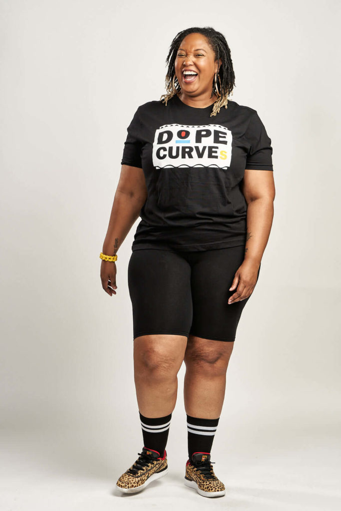 Black woman laughing wears black biker shirts and black shirt that says “Dope Curves” and leopard print sneakers