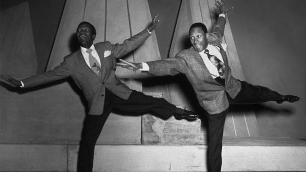 African American dancers in suits black and white with their arms outstretched on one leg