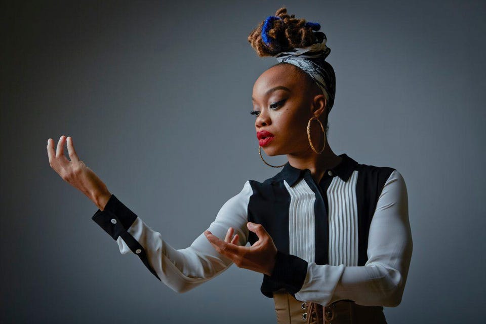 Camille A. Brown choreographer wears looks at her outstretched hands wearing large hoops, red lipstick and a traditional hairstyle in front of a gray background 