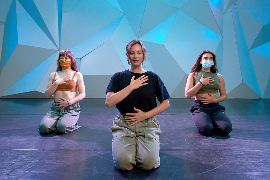 CLI Instructor Mackenzie Dustman kneels on the ground with two other dancers with their hands on their heart and stomach 
