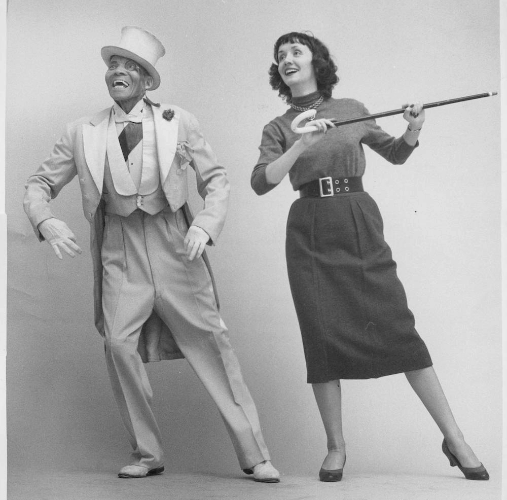 Black and white photo of a white woman with a cane and an African American man with a top hat and a monocle smiling together looking out in the distance