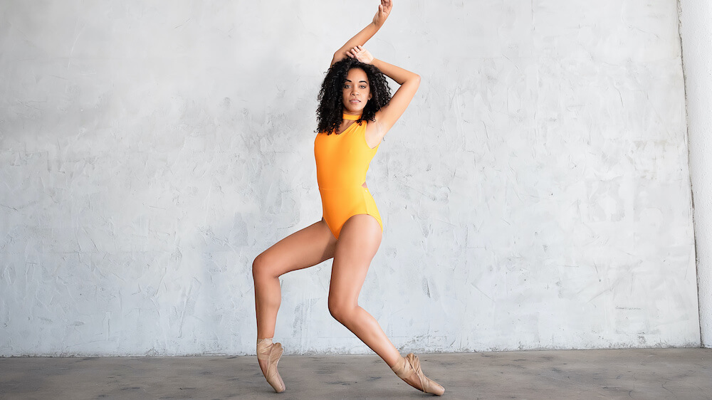 Dancer in pointe shoes wearing a yellow leotard with brown curly hair standing in front of a grey wall with her legs bent and her arms over her head