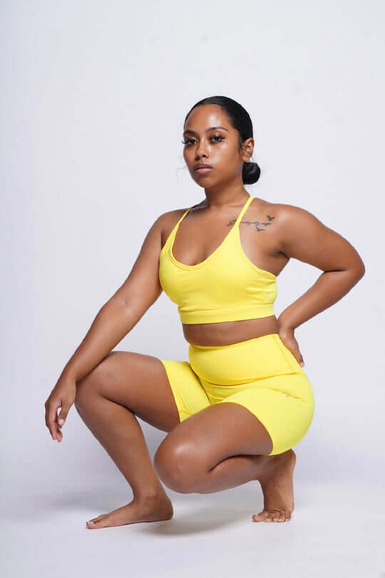 Woman in yellow sports bra and biker shorts matching set squatting with her hand on her back gazing at the camera