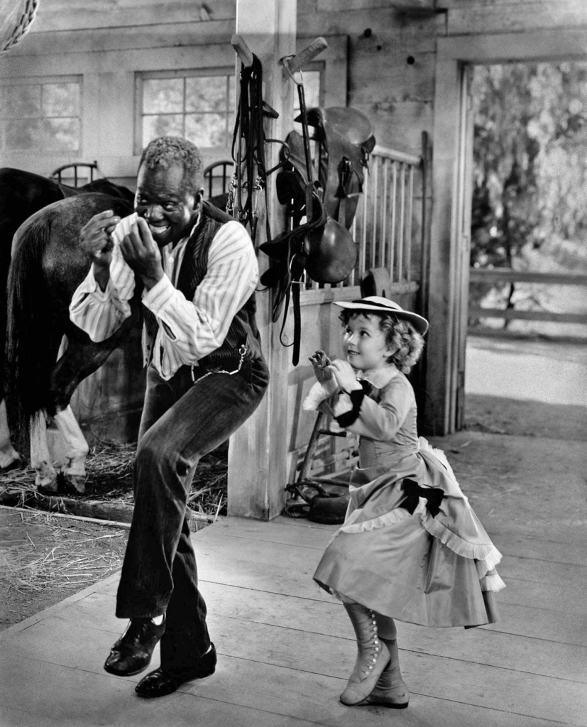 Black and white of Bill Robinson, known as “Bojangles,” dancing and smiling with Shirley Temple in a barn