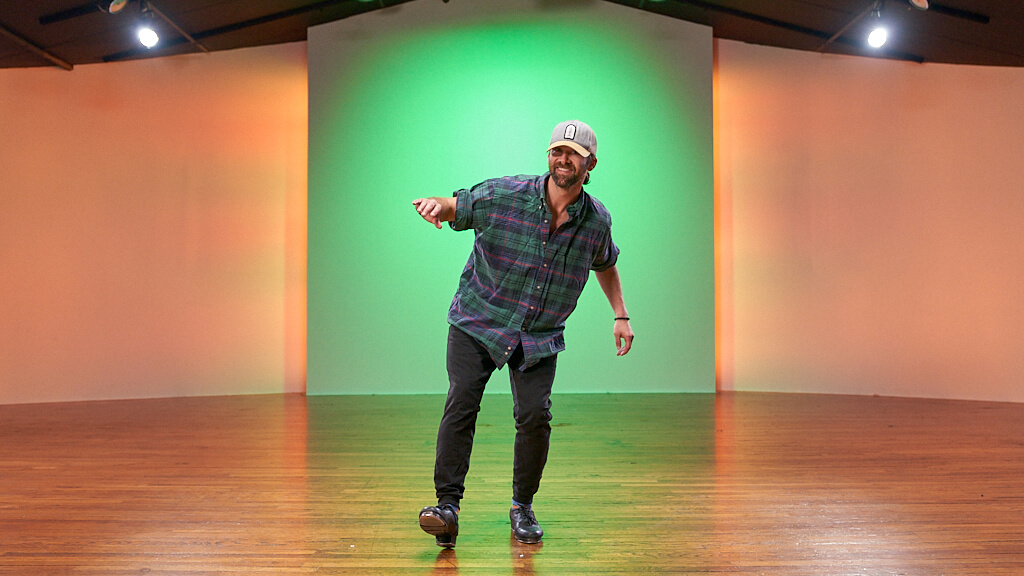 Mike Keefe tap dancing in CLI Studios in front of a green and orange background on wooden dance floor wearing a flannel