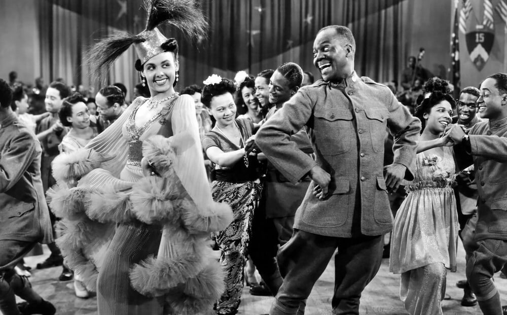 Bill “Bojangles” Robinson with woman in feathered hat and fancy dress, smiling and dancing in a crowd, black and white