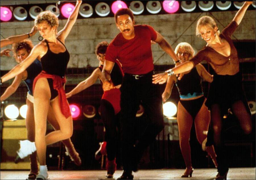 Gregory Hines in red shirt and black pants dancing with women in leotards  80s style in a club setting