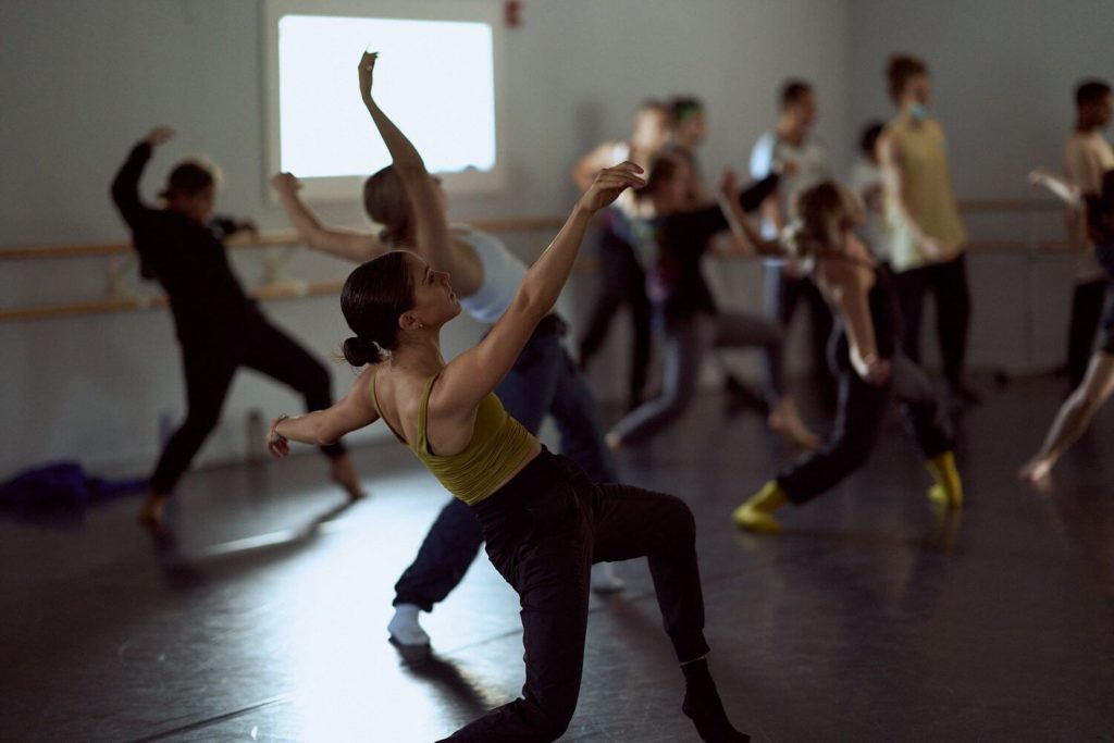 Dancer leaning back off center with one leg bent at the CLI Conservatory with dancers in the background of dance studio
