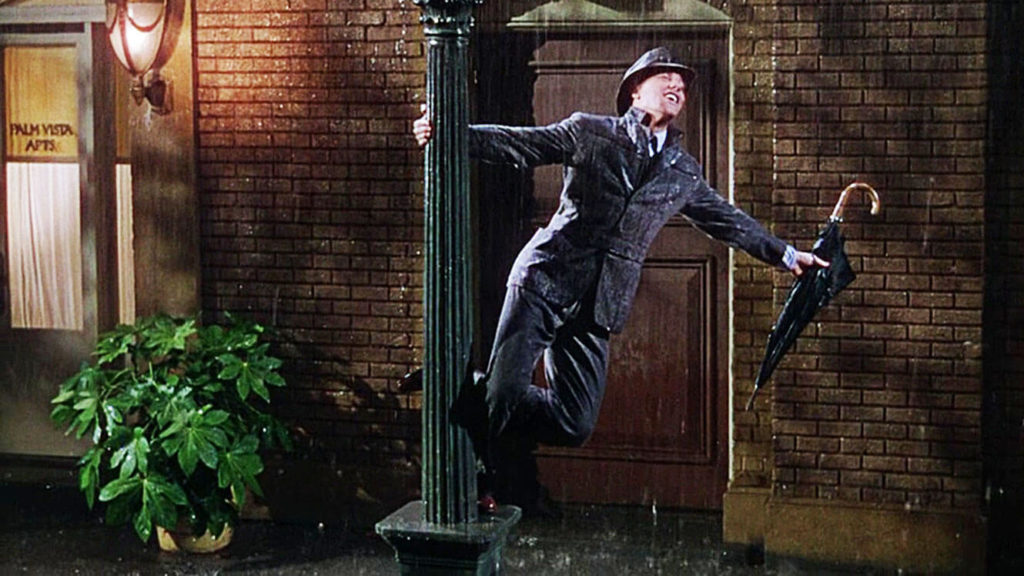 Gene Kelly wearing suit and hat with umbrella in hand swinging off a lamppost in Singing in the Rain in front of a wet brick house 