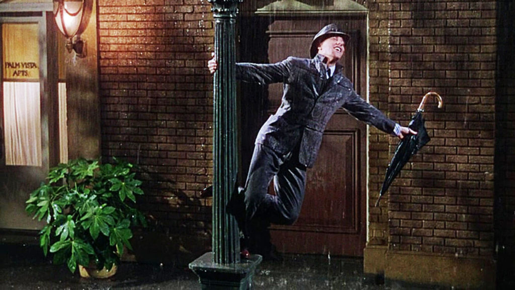 Gene Kelly wearing suit and hat with umbrella in hand swinging off a lamppost in Singing in the Rain in front of a wet brick house